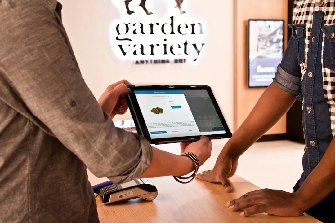 A budtender and cannabis customer discuss the merits of various cultivars in a dispensary. Between them is a lit tablet displaying Covas point of sale cannabis software that they are using to review the customers order options together.